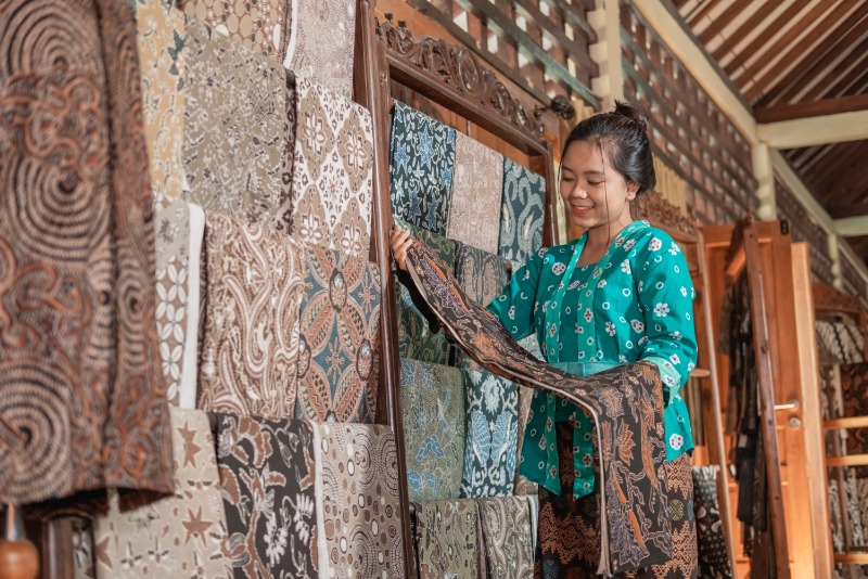 Seorang pengrajin di Indonesia sedang fokus menerapkan lilin malam dengan canting pada kain batik, menunjukkan keahlian dalam membuat motif tradisional yang kompleks