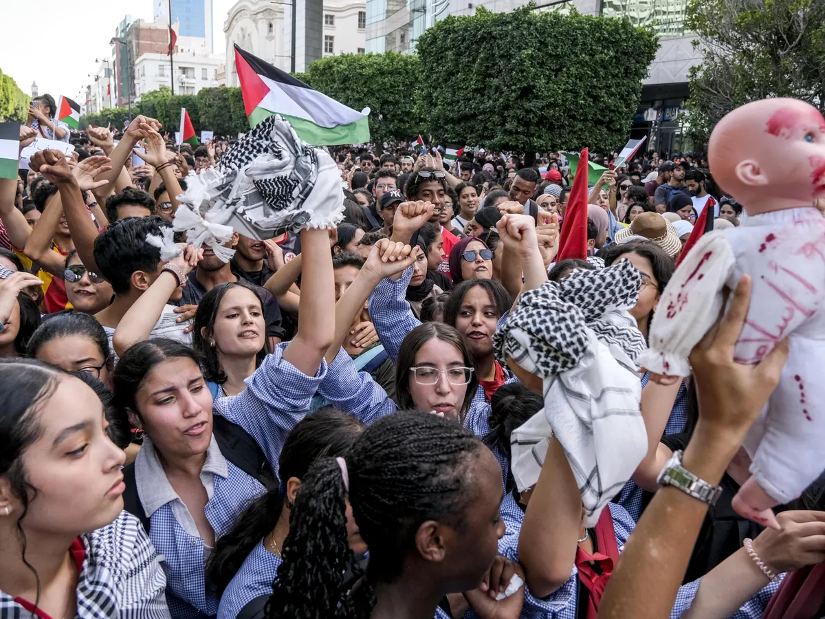 A group of people holding signs and flags protesting in a city street against government suppression and in support of Palestinian rights, with banners displaying messages of solidarity and freedom.